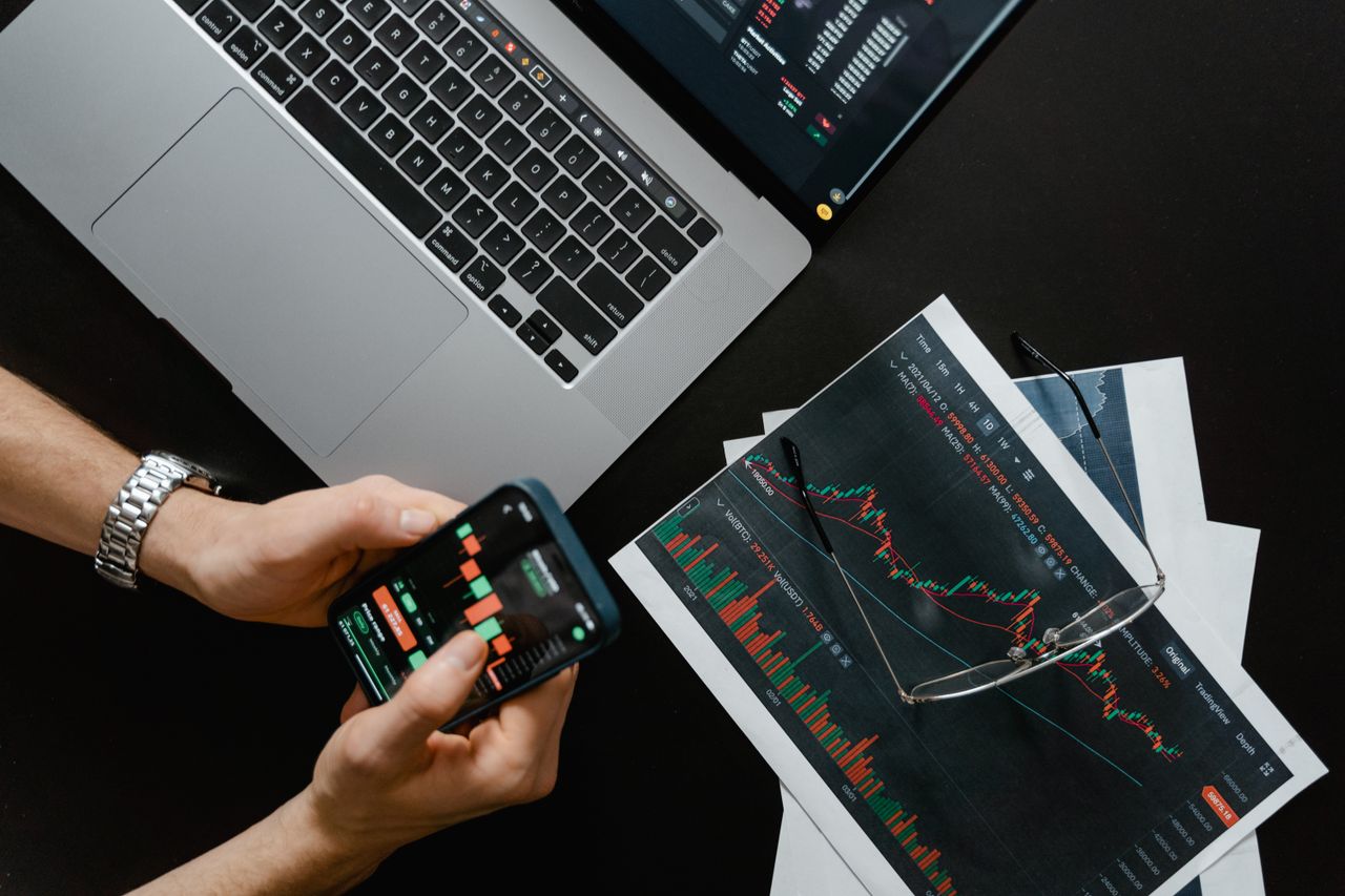 why-choose-us Hands using smartphone beside laptop with stock charts, showcasing digital trading.